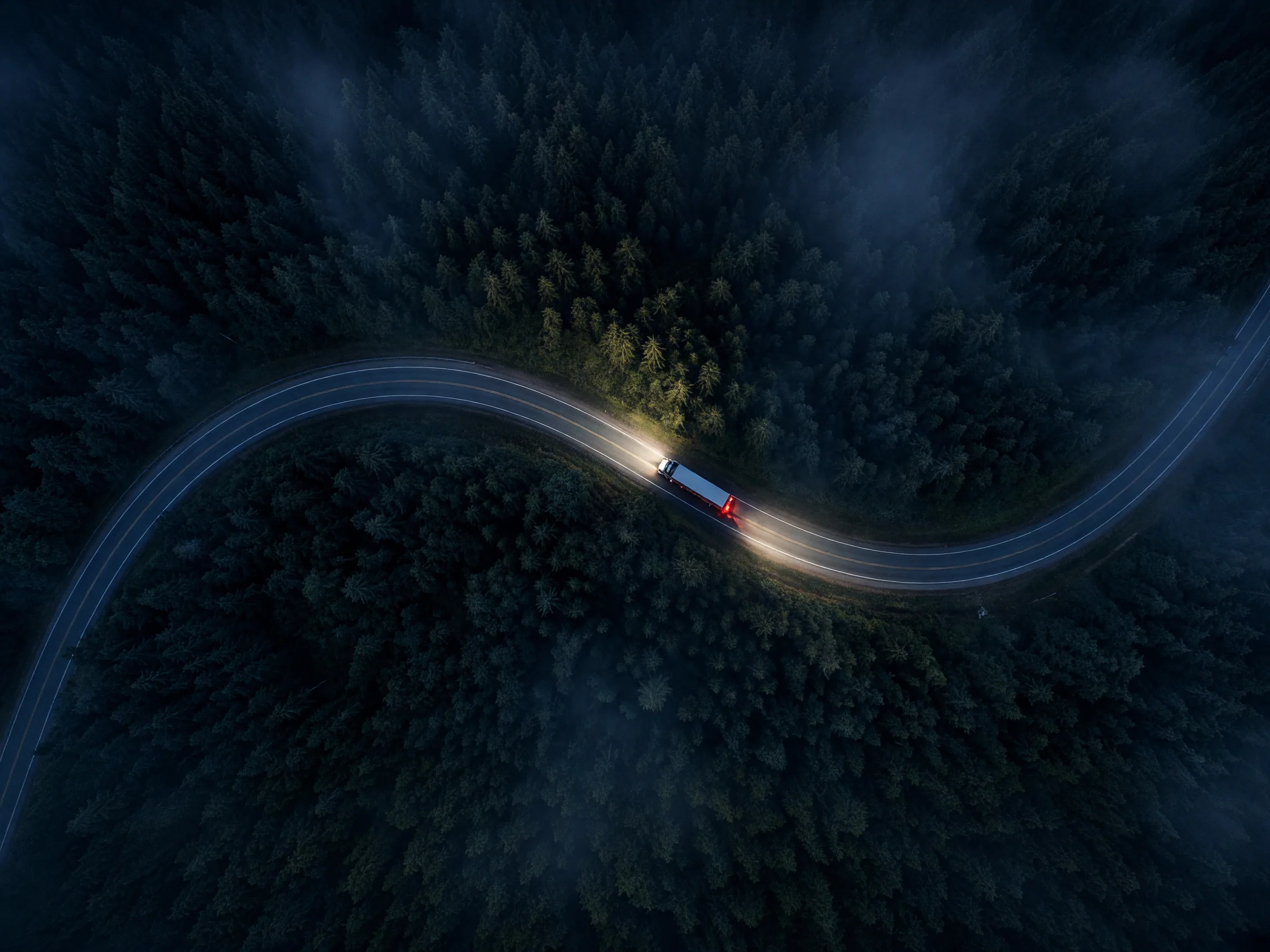 Aerial night view of a semi truck running with headlights on down a curving two-lane highway through dense evergreen forest